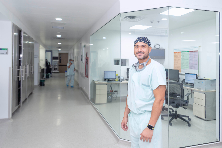 En un pasillo de hospital se ven en primer plano al lado derecho una oficina en las cuales sus paredes son de cristal, frente a esta oficina hay un médico mirando a la cámara.