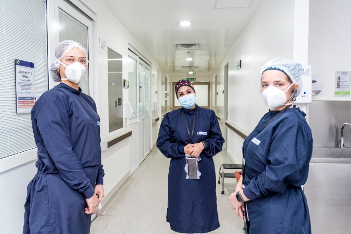 En un pasillo de hospital se ve a tres profesionales de la salud mirando hacía al frente todas con traje y gorro quirúrgico y tapabocas.