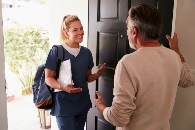 En el umbral de la puerta de una casa hay dos personas, en la parte interna de la casa hay un hombre y en la parte externa hay una mujer que por su vestimenta se puede inferir que es funcionaria del sector salud, ella tiene una cara amable y parece estar explicando algo
