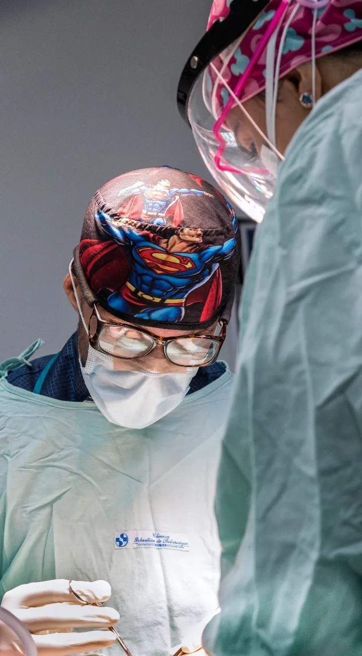 Hay dos médicos mirando fijamente lo que se infiere como un campo quirúrgico, al de la derecha solo se le ve el brazo y una mínima parte del rostro y al que está de frente se le ve un gorro quirúrgico de supermán y unas gafas. 