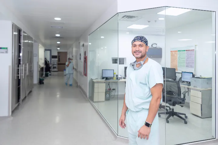 En un pasillo de hospital se ven en primer plano al lado derecho una oficina en las cuales sus paredes son de cristal, frente a esta oficina hay un médico mirando a la cámara