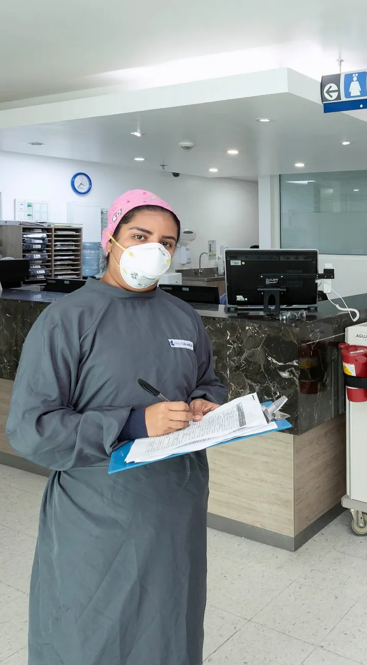 Frente a lo que se infiere como una estación de enfermería se encuentra a una profesional de la salud usando cubrebocas, gorro quirúrgico, mirando hacía al frente y sosteniendo una planilla y un lapicero. 